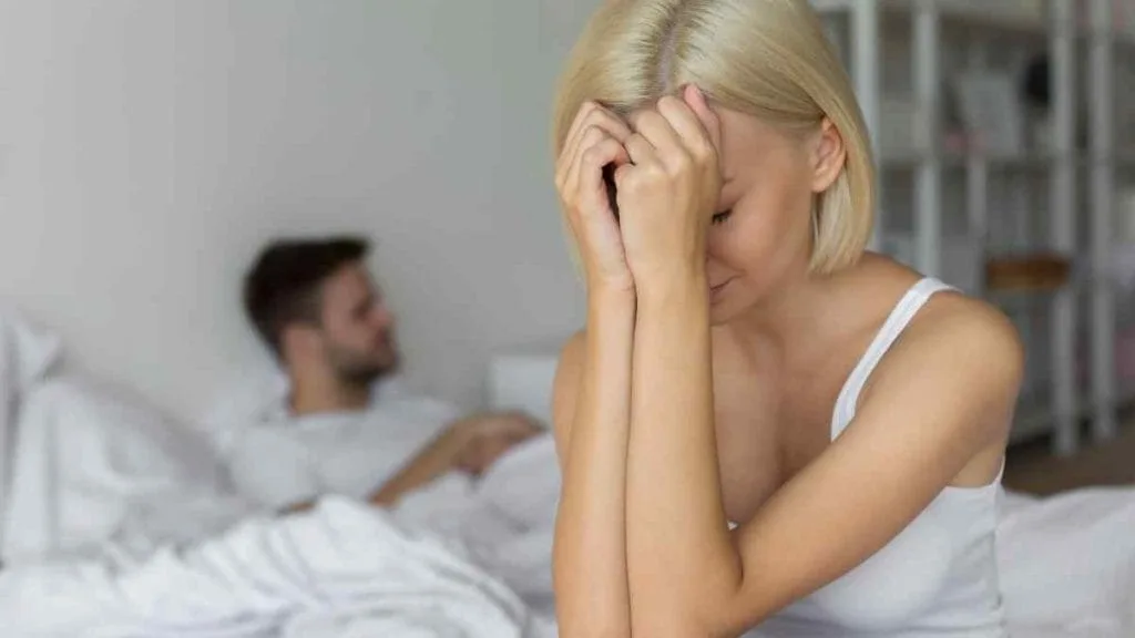 Mulher sentada na cama com a mão no rosto, e um homem deitado ao fundo
