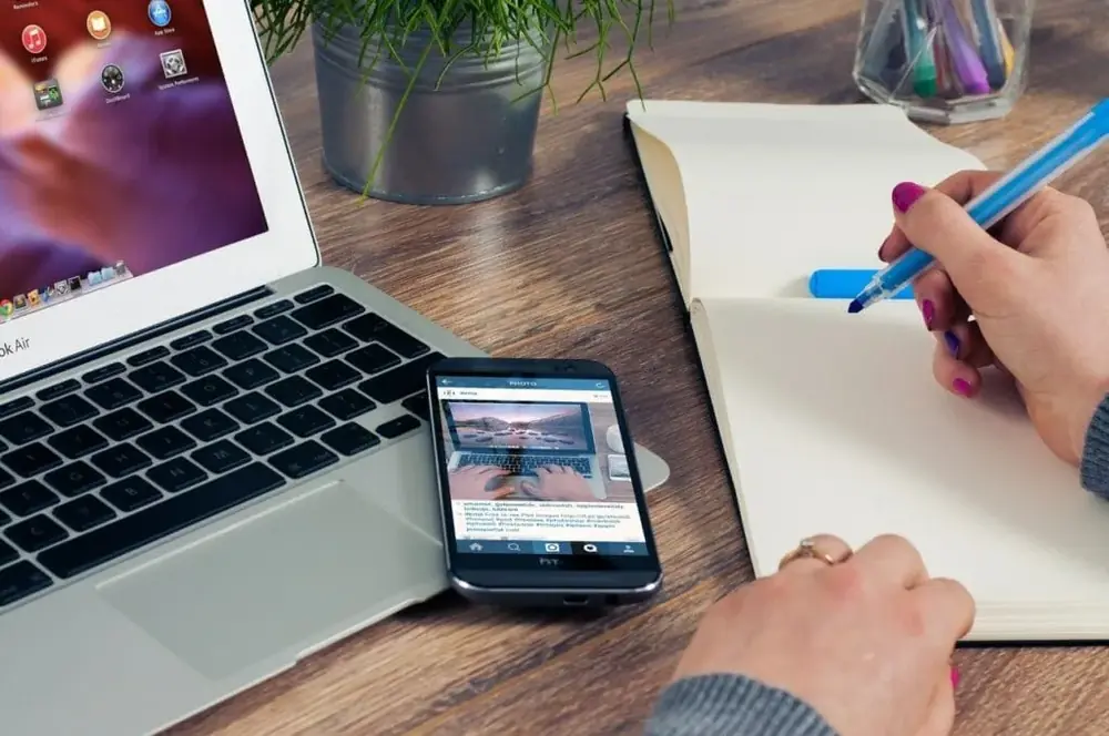 Mesa com notebook, celular e uma mão escrevendo num caderno