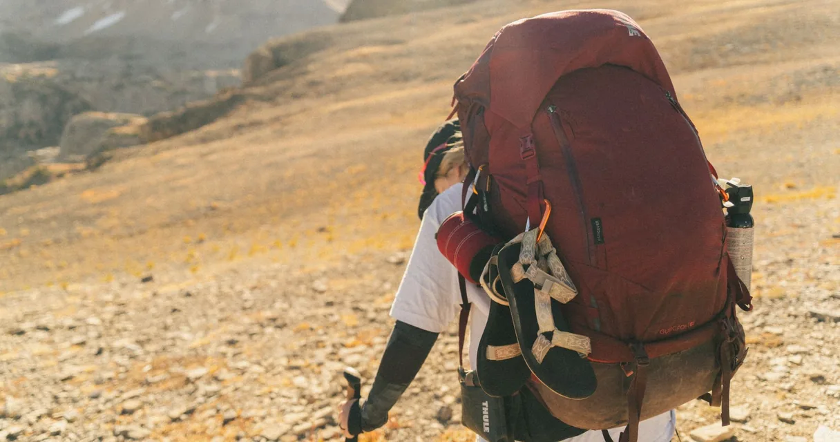 Pessoa carregando uma mochila grande e pesada nas duas alças em uma trilha de montanha